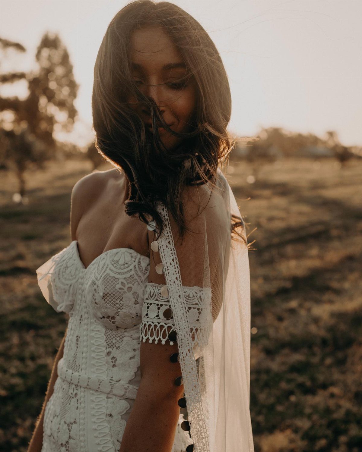 Boho Wedding Dress Looks: 8 Ideas from Lace to Chiffon 6 Woman in a white lace boho wedding dress with a sheer cape in a field.