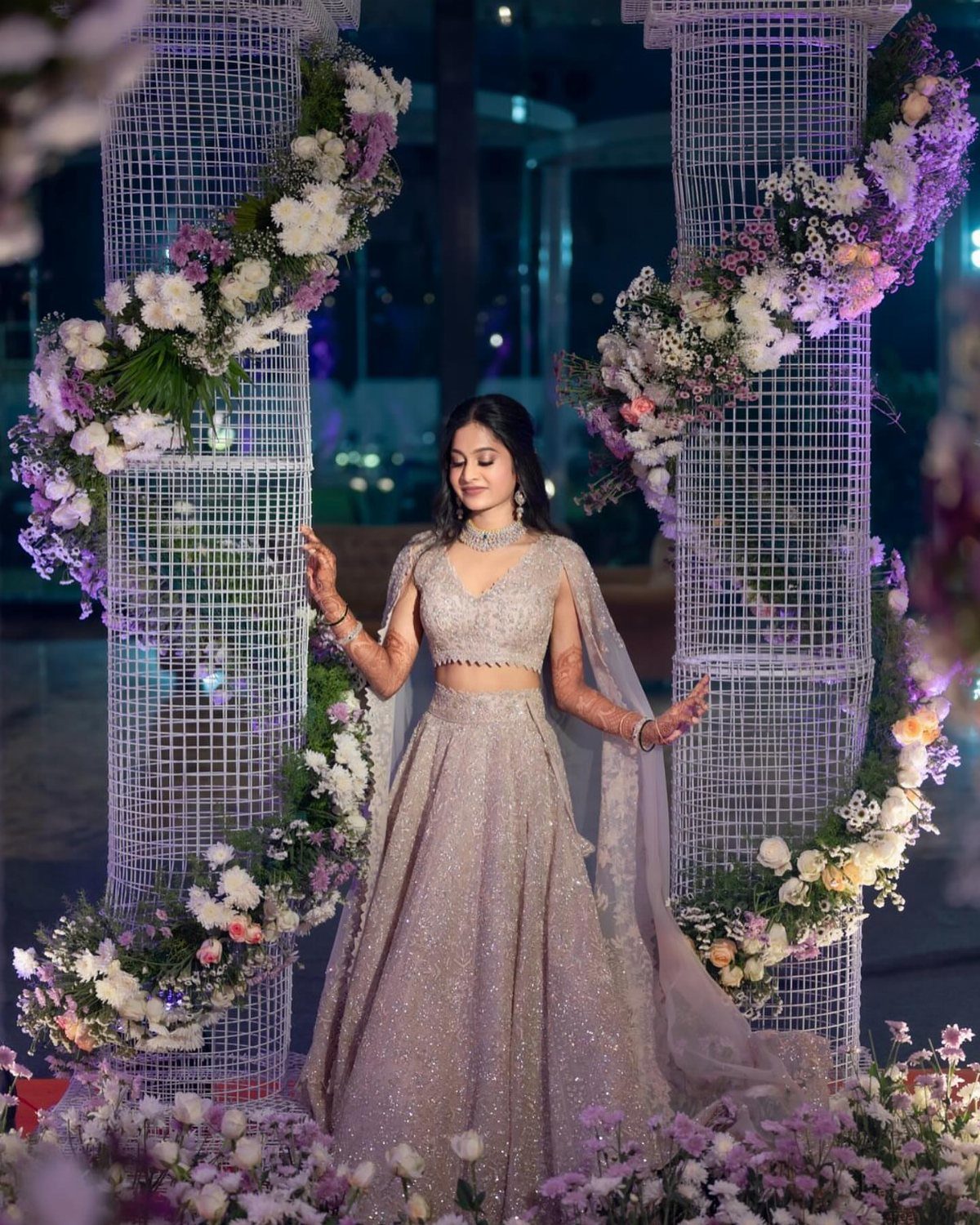 Bride Reception Dress: 17 Ideas for Your Second Look 16 Woman in a shimmering purple lehenga standing between floral pillars at a reception.