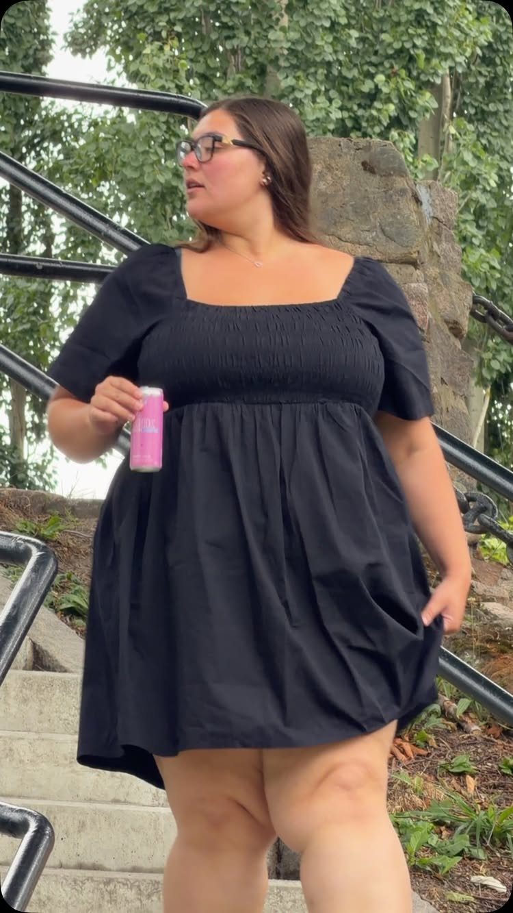 Woman wearing a short black smocked summer dress standing on outdoor concrete stairs.