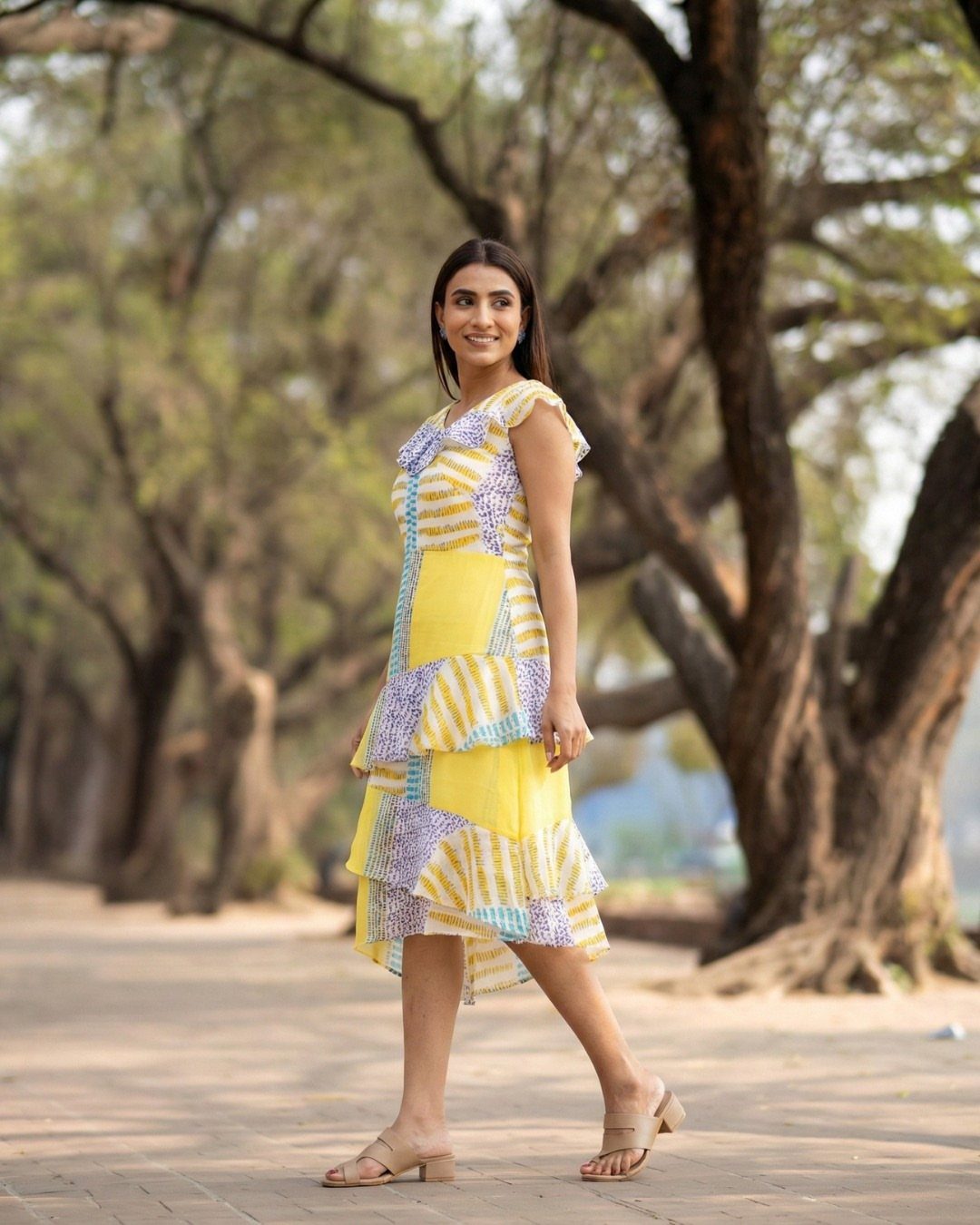 Woman wearing a yellow and purple patterned midi dress walking on a tree-lined path.