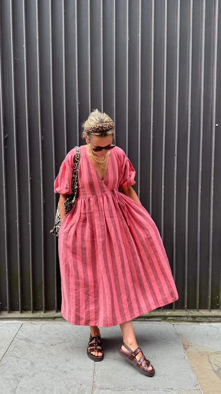 Woman in a pink striped summer midi dress and brown sandals against a grey wall.