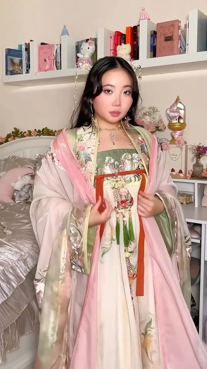 Woman wearing a traditional pink and green Hanfu with floral embroidery in a bedroom.