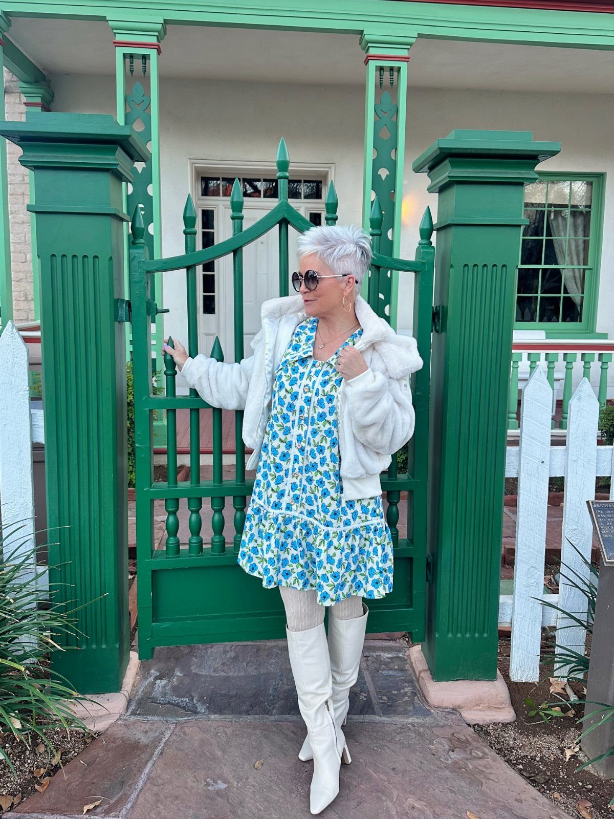 Classy Winter Dresses in 14 Timeless Styles 10 Woman in floral dress, white jacket, and white boots by a green gate.