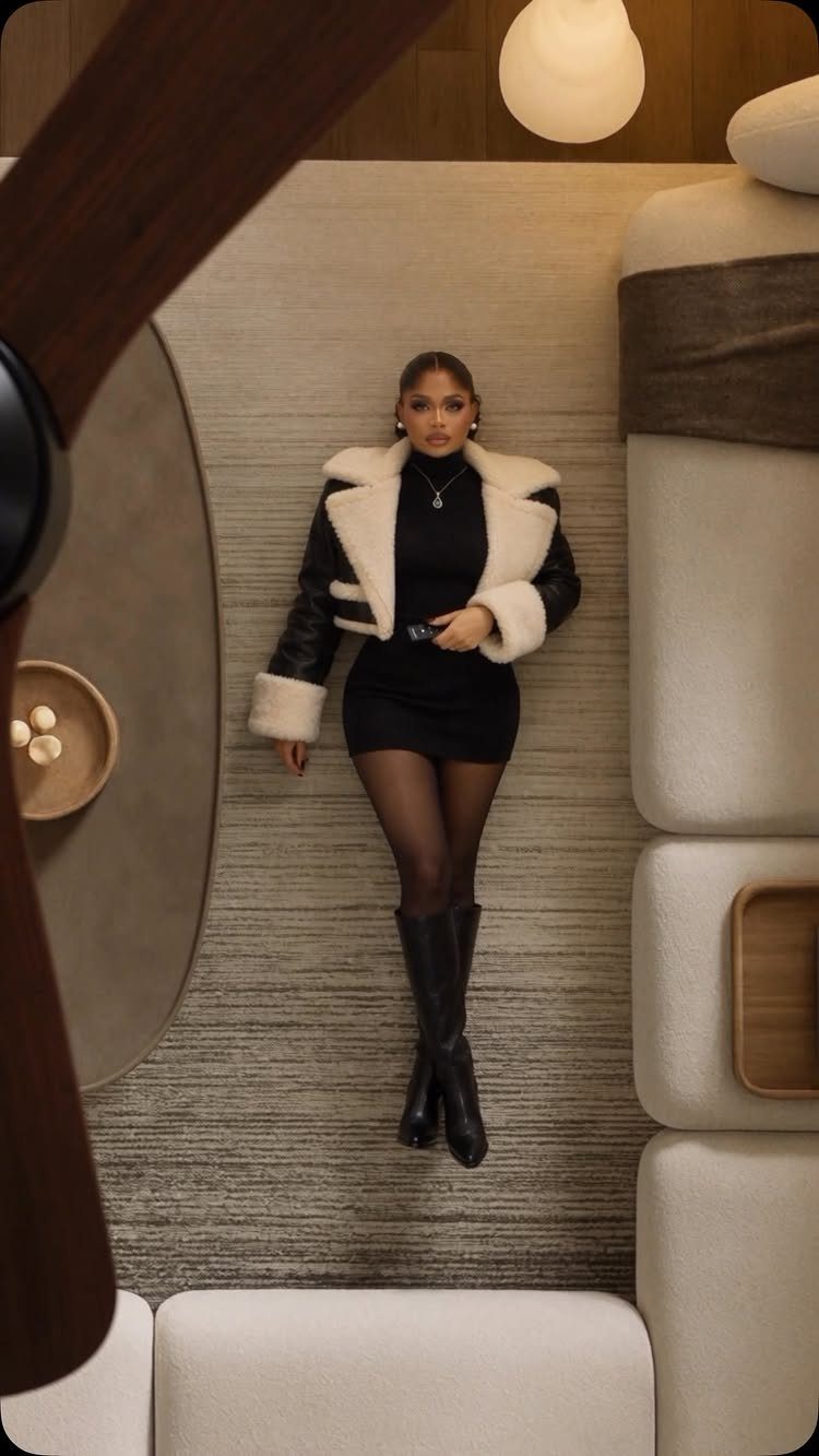 Classy Winter Dresses in 14 Timeless Styles 13 Woman in a short black dress and shearling jacket standing in a modern room.