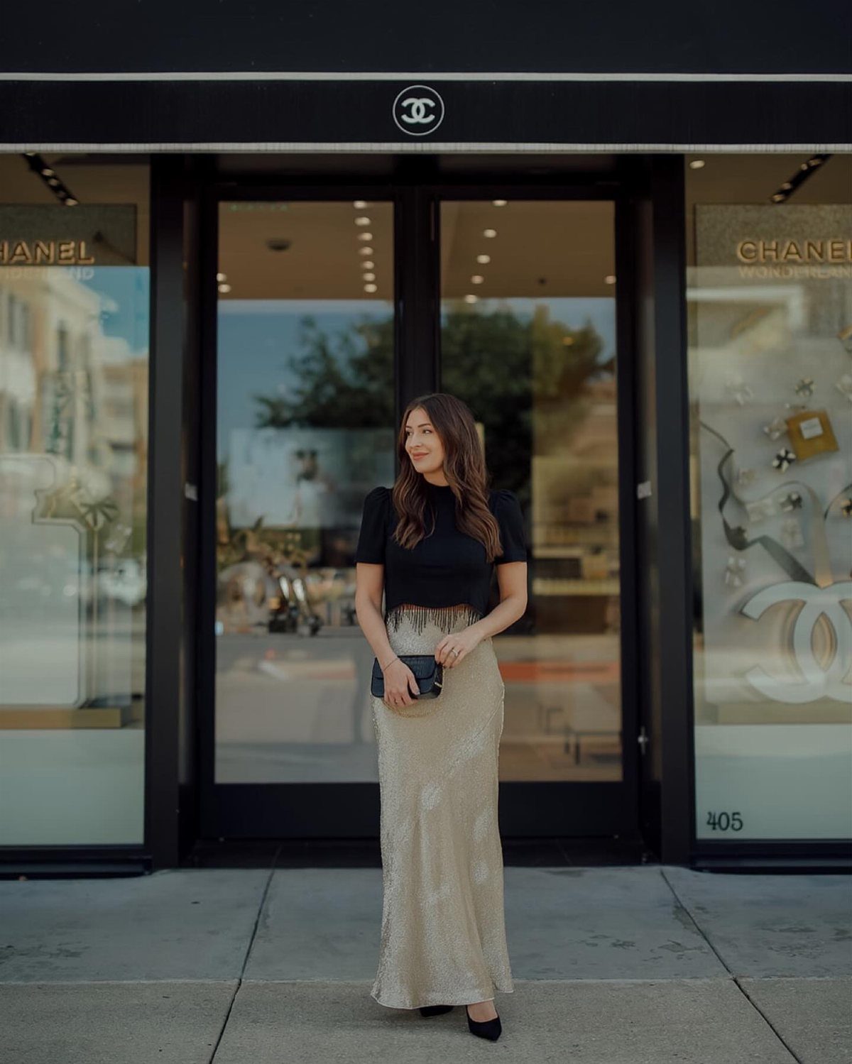 Woman wearing a black top and long shimmering gold sequined skirt outside a store.