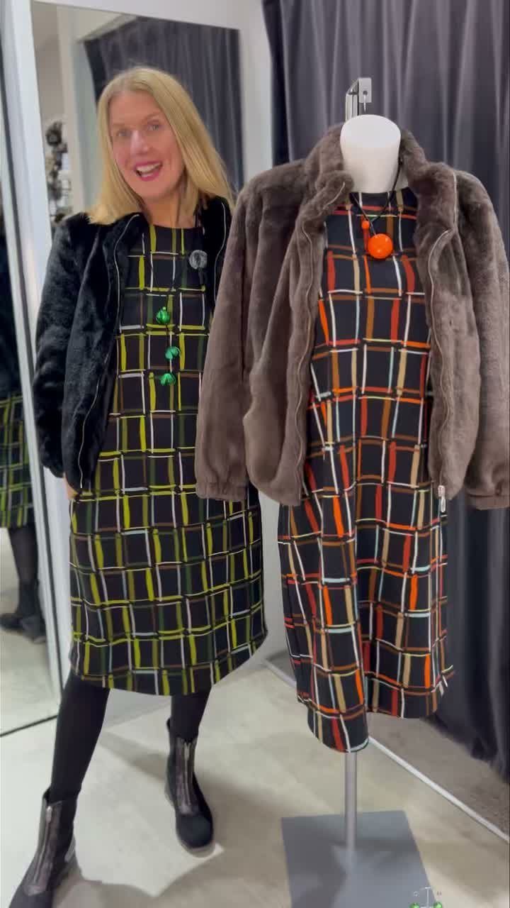 Woman and mannequin wearing faux-fur jackets over plaid dresses in a clothing store.