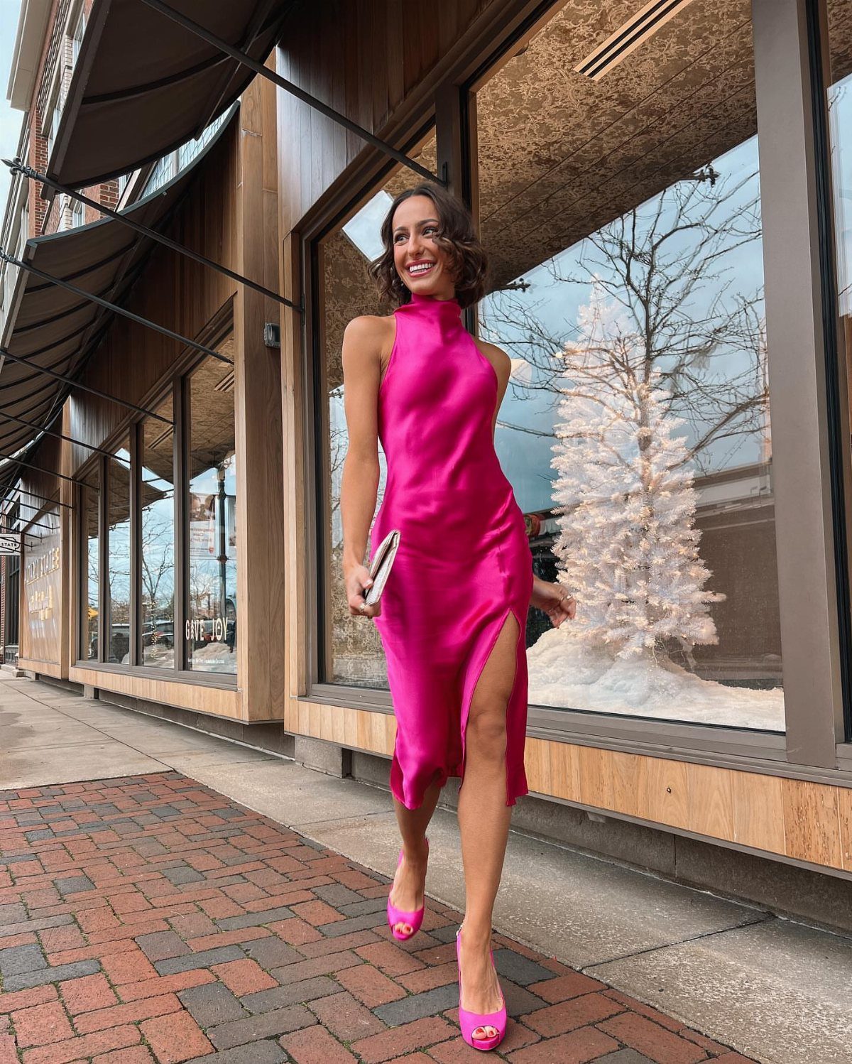 Woman in a bright pink satin midi dress standing by a storefront Christmas tree.