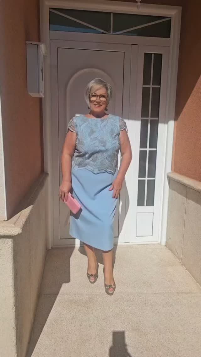 Older woman wearing a light blue midi dress and silver heels standing by a door.