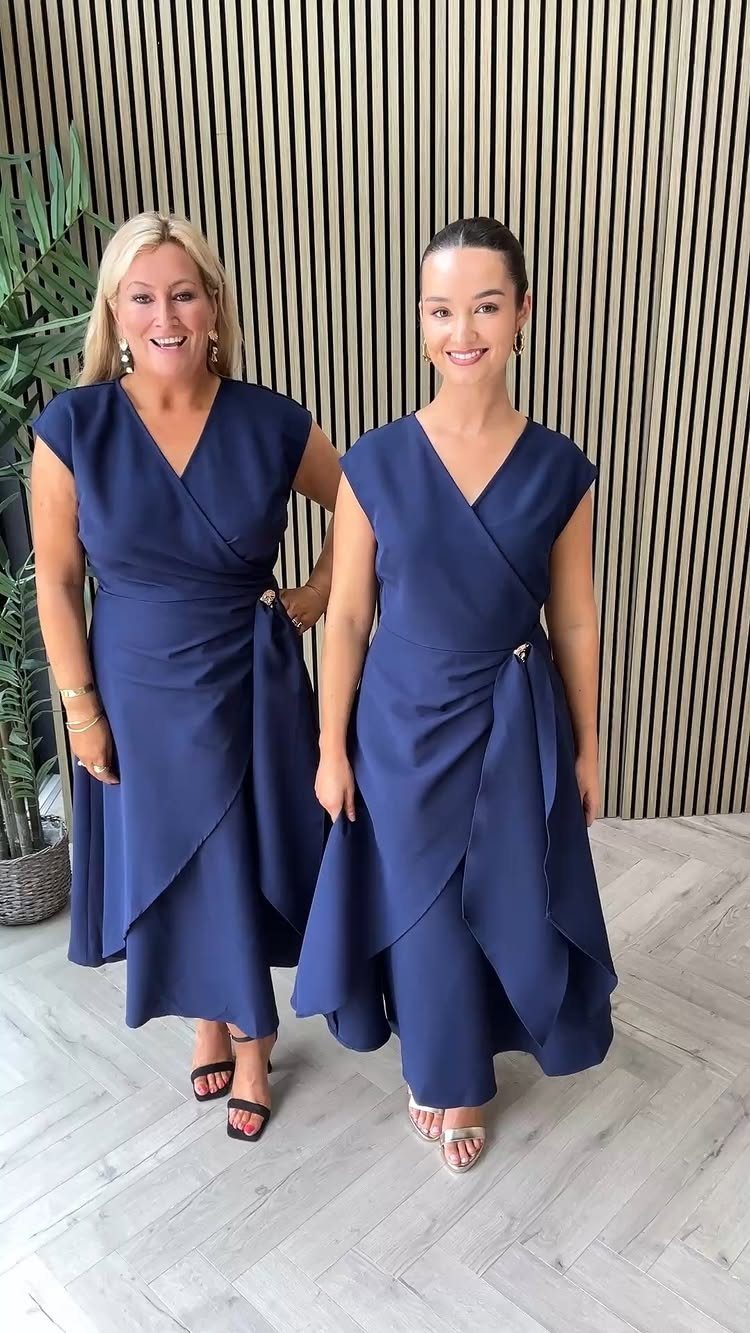 Two women wearing navy blue wrap dresses standing against a wooden slatted wall.
