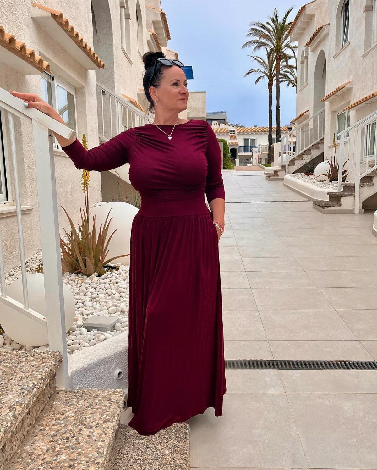 Woman in a long-sleeved burgundy maxi dress standing on a sunny outdoor walkway.