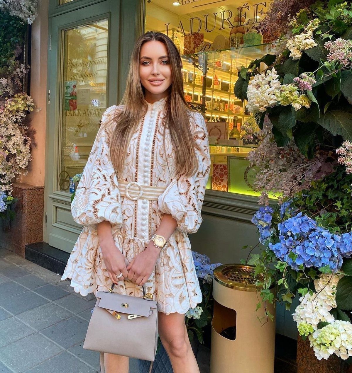 Woman in a short cream textured dress with a belt and beige handbag.