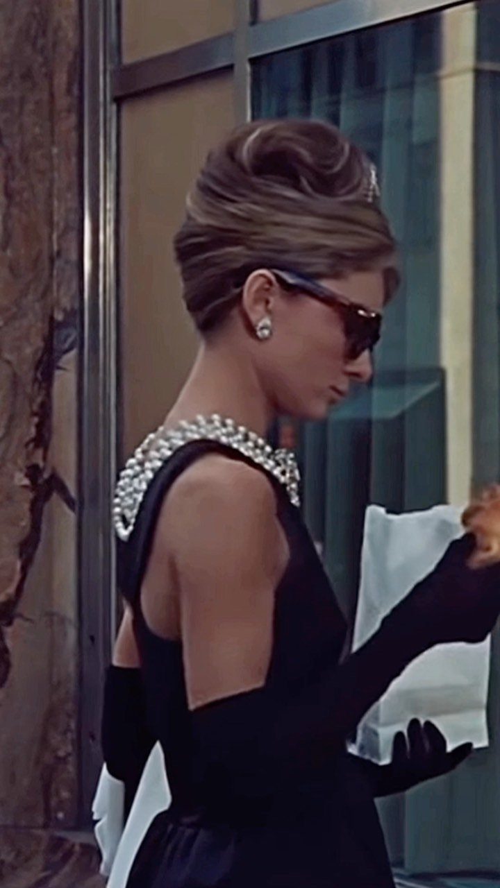 Woman in a black dress, pearls, and sunglasses with a high updo hairstyle.