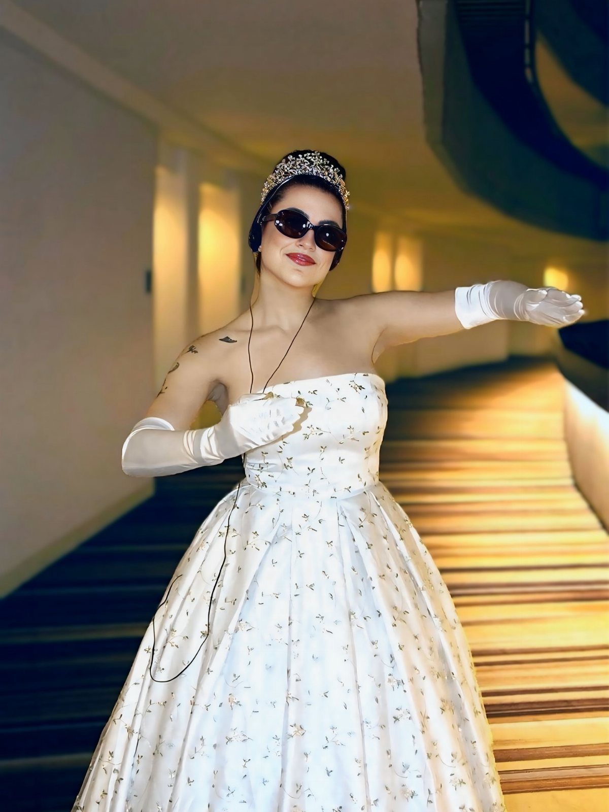 Woman in a white floral ballgown, tiara, and sunglasses posing in a hallway.