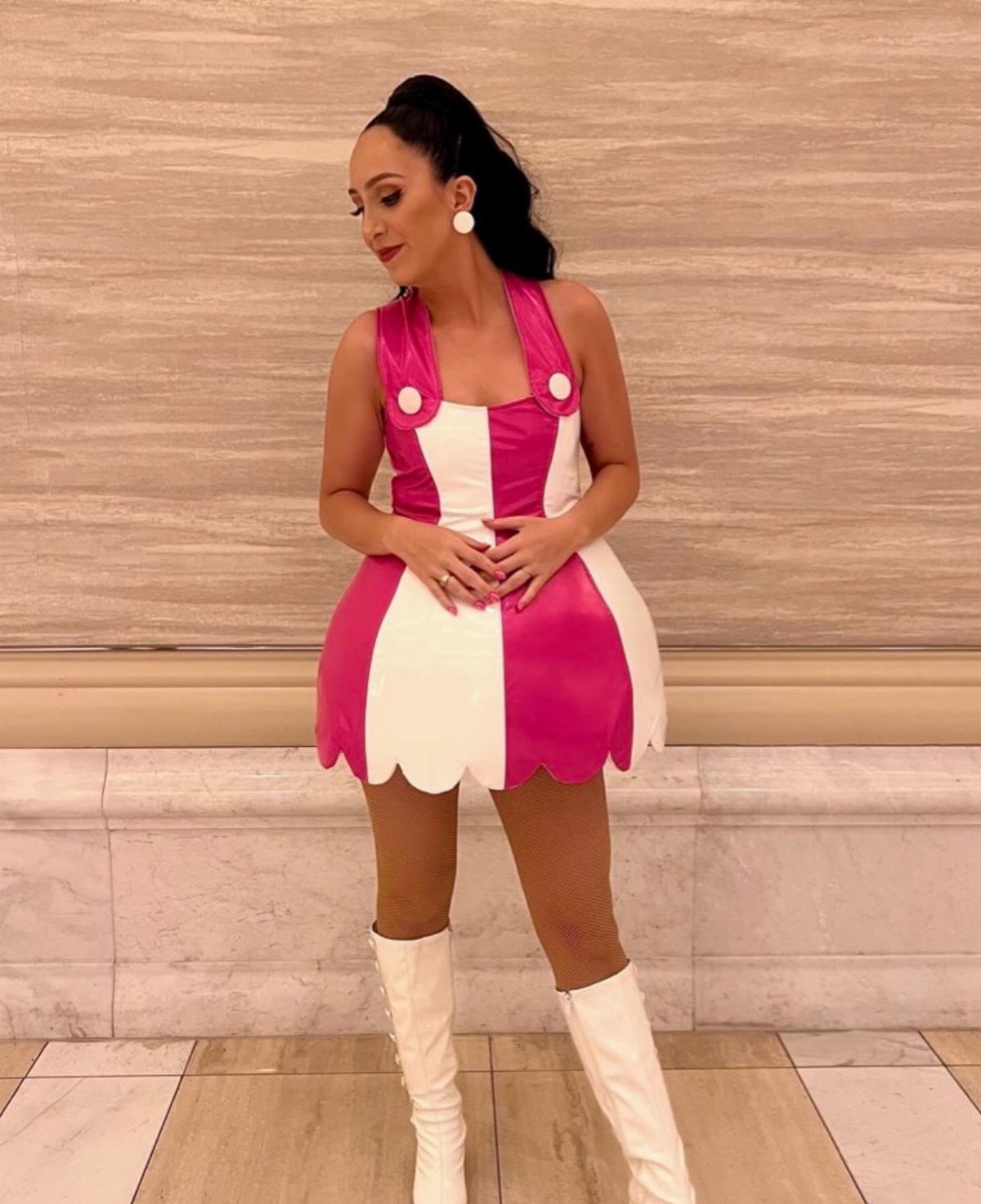 Woman in a pink and white striped dress and white boots posing indoors.