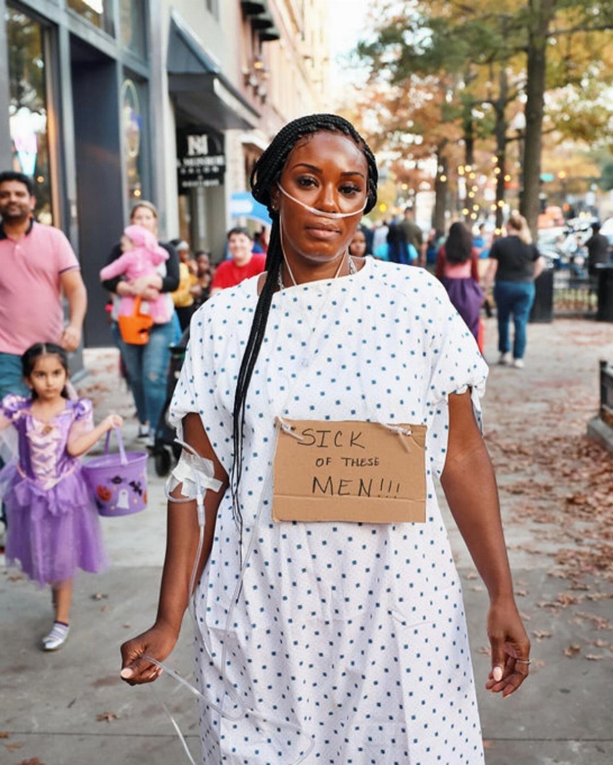 Woman in a hospital gown and nasal cannula holding a sign on a sidewalk.