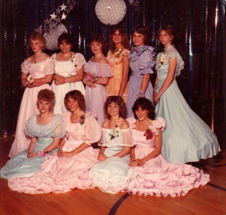 Ten young women wearing colorful, ruffled eighties prom dresses posing in a gymnasium.