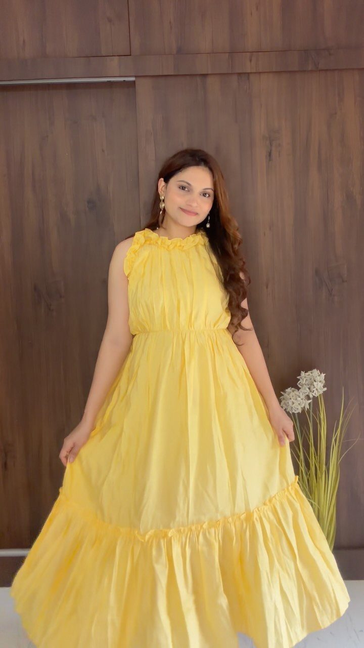 Woman in a flowing yellow sleeveless maxi dress posing against a wooden wall.