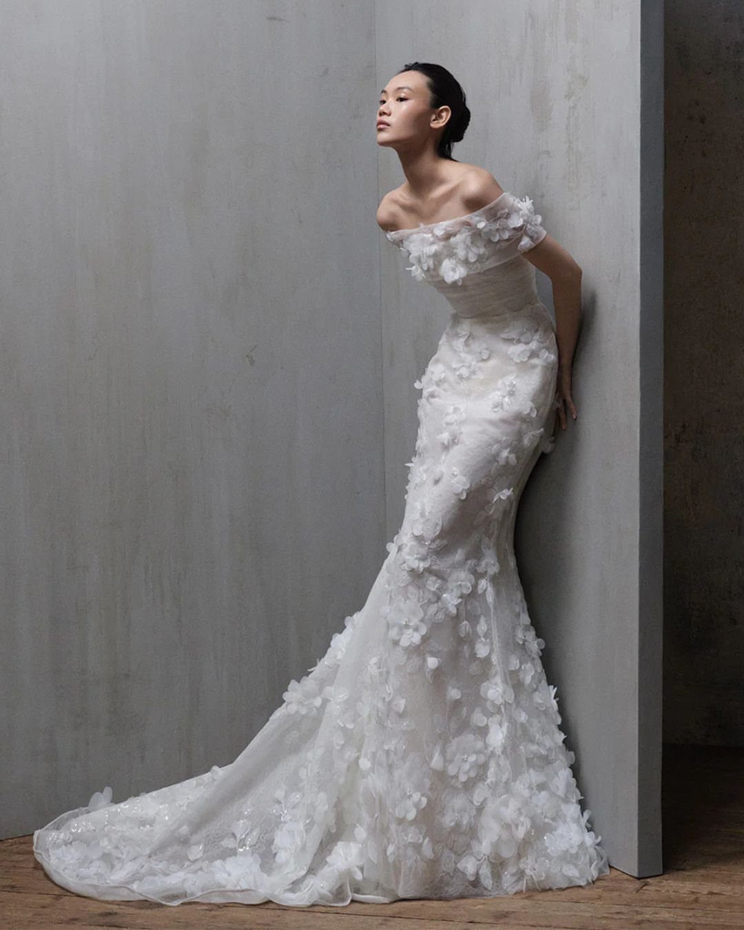 Woman in a white off-the-shoulder floral mermaid gown leaning against a grey wall