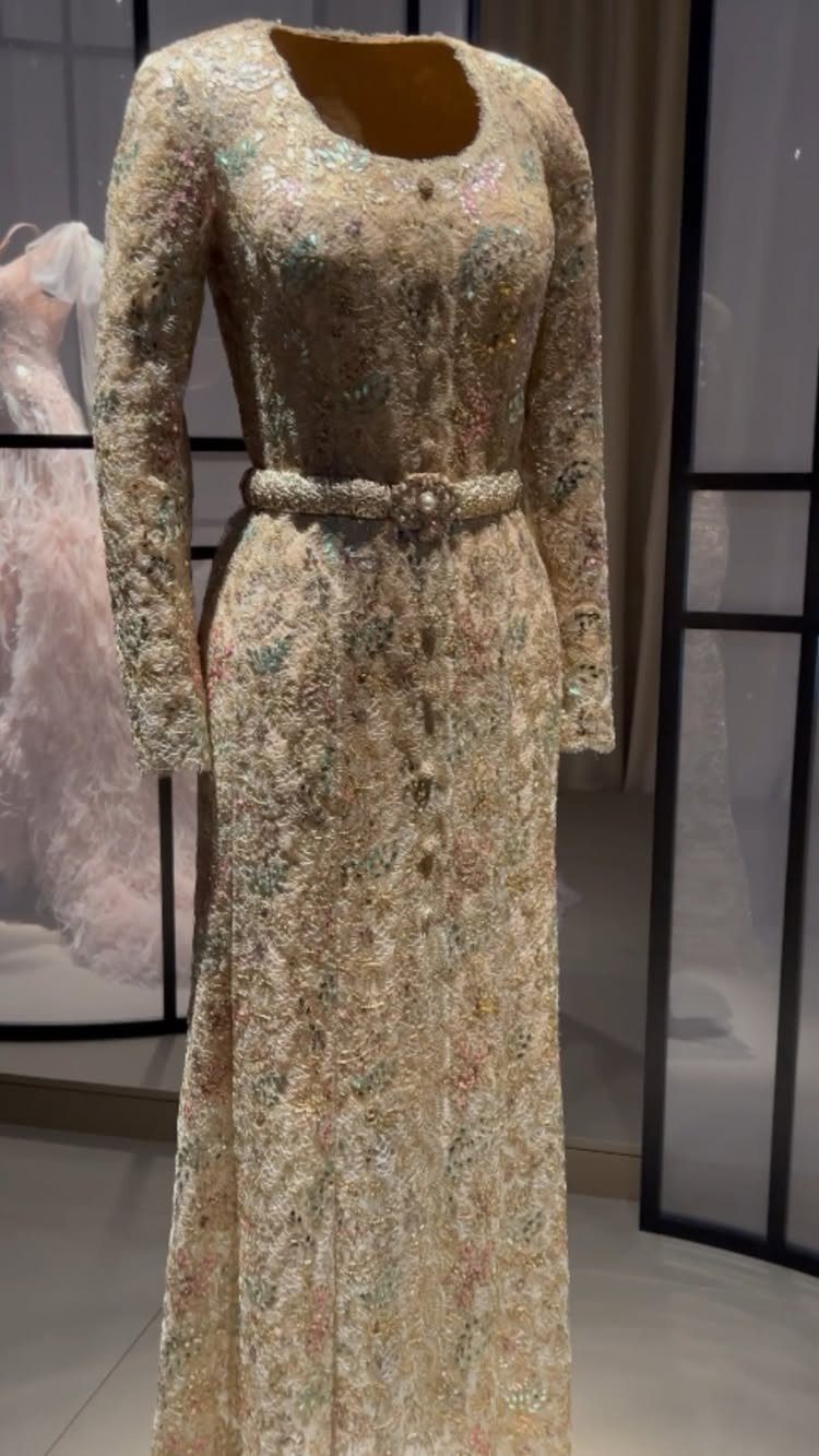 Intricately beaded champagne long-sleeved gown with a floral belt on a mannequin.