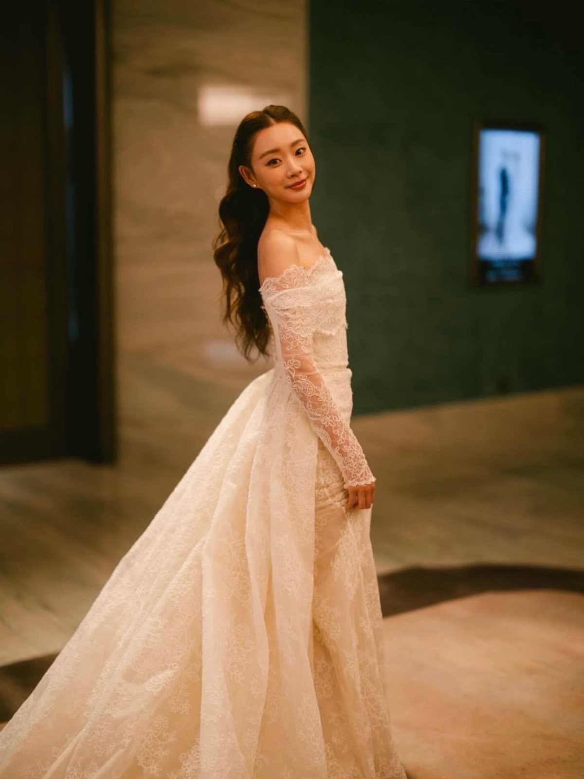Woman in a voluminous cream lace off-the-shoulder gown standing in a modern interior.