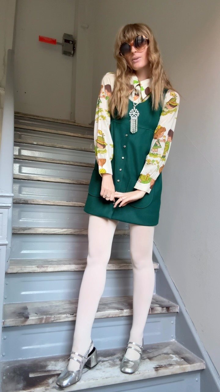 Woman wearing a green jumper dress with a patterned blouse and silver shoes.