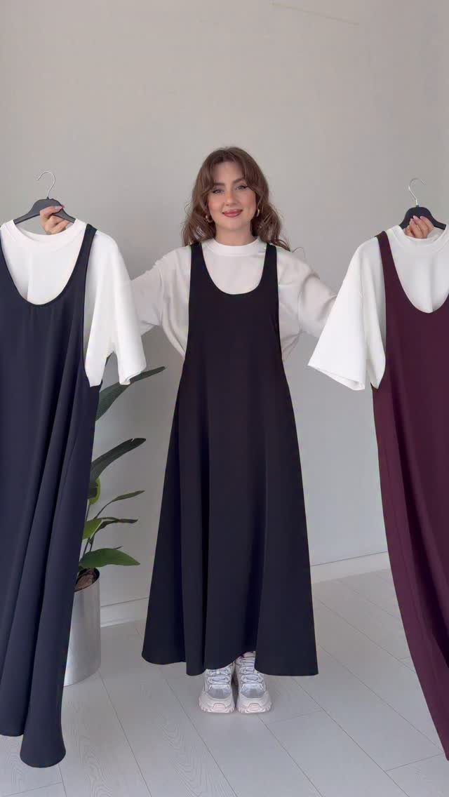 Woman wearing a black jumper dress over a white shirt holding other dress options.
