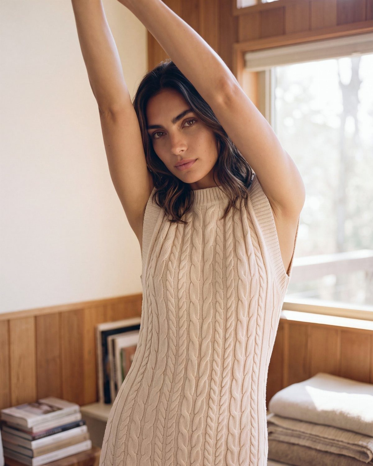 Woman wearing a cream cable-knit sleeveless jumper dress posing in a sunlit wooden room.