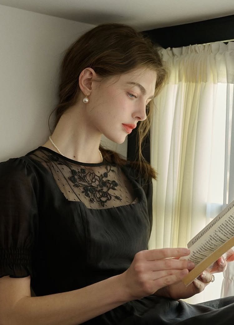 Woman in a black short-sleeved dress with lace detailing reading a book indoors.