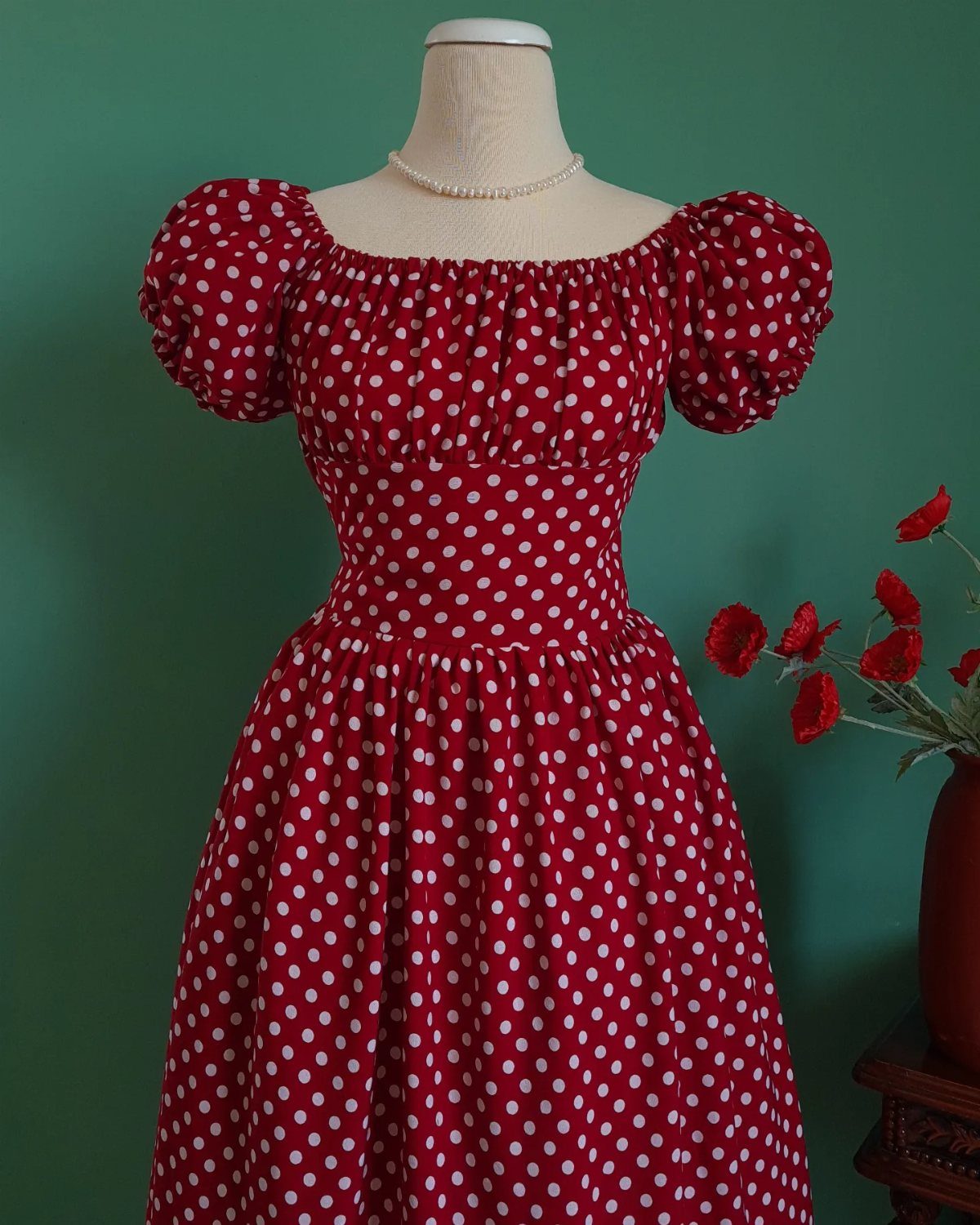 Red polka dot dress with puffed sleeves on a mannequin with pearl necklace.