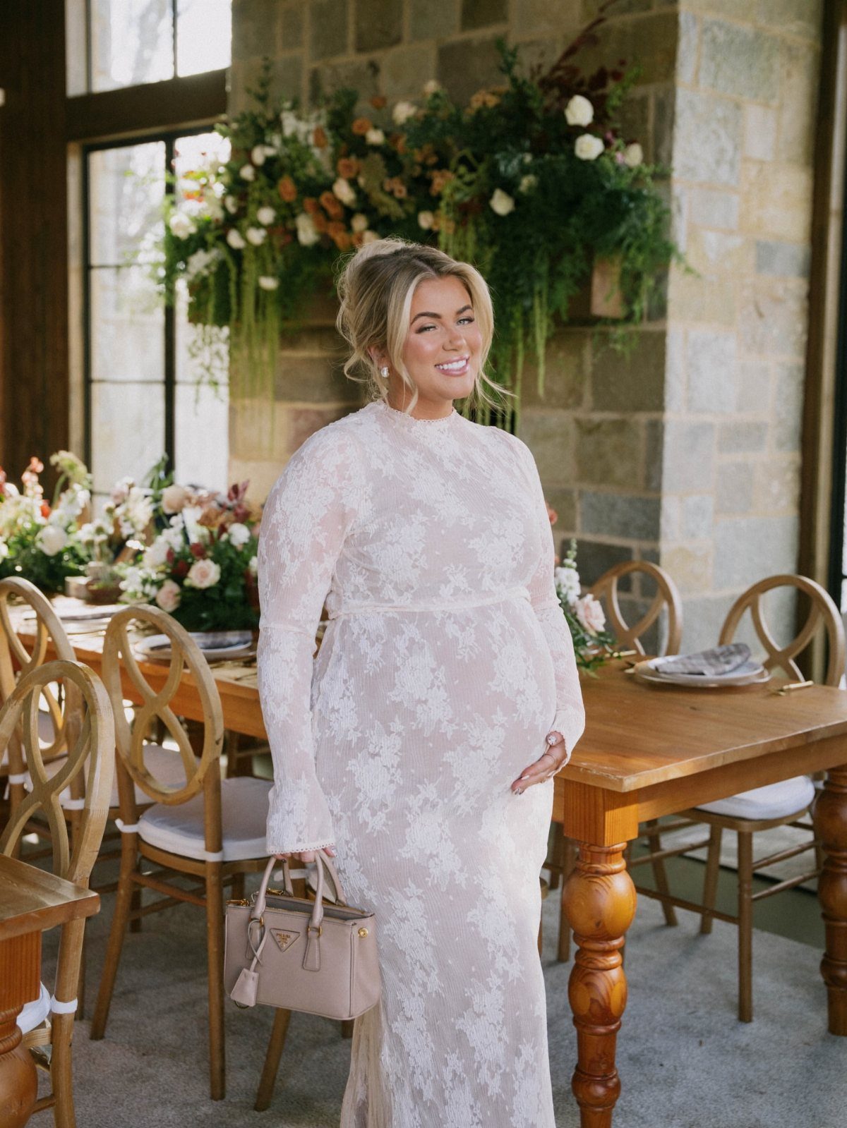 Pregnant woman in a white lace maternity dress standing by a decorated wooden table.