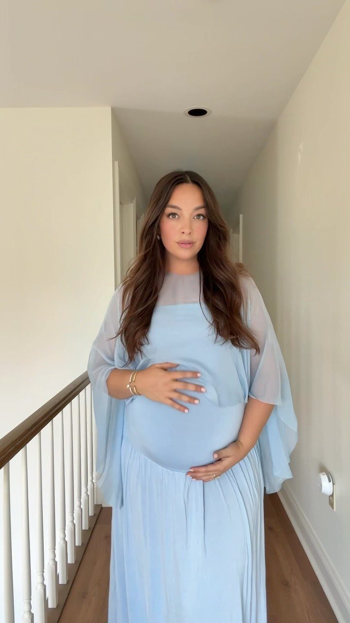 Pregnant woman in a light blue maternity dress standing in a bright hallway.