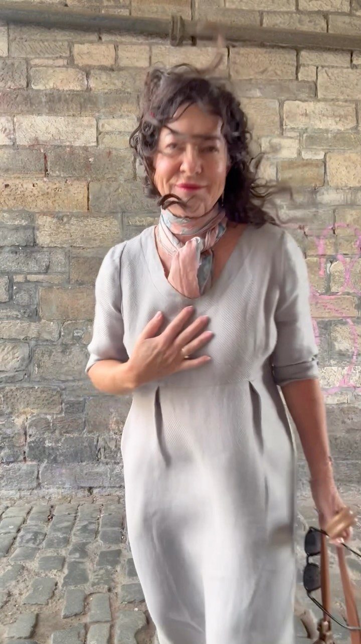 Woman in a grey midi casual dress and pink scarf standing by stone wall