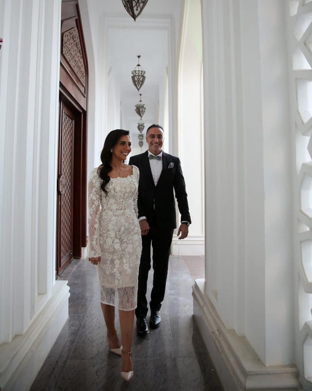 Woman in a white floral lace midi dress walking with a man in tuxedo.