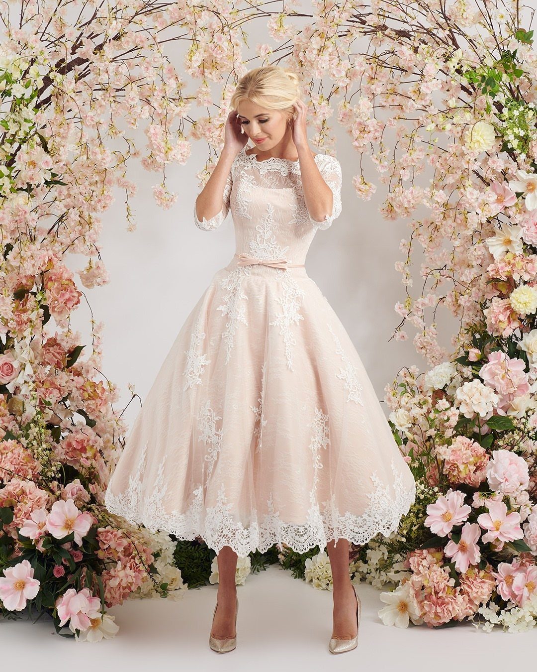 Woman in a pale pink tea-length lace wedding dress standing among cherry blossoms.