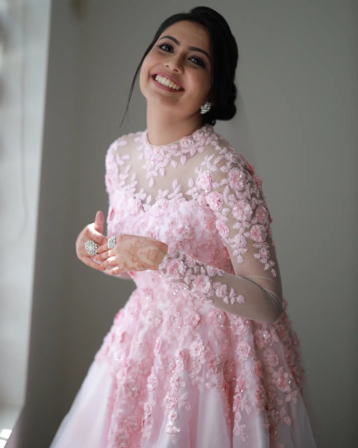 Smiling woman in a light pink long-sleeved gown with floral lace appliqués.
