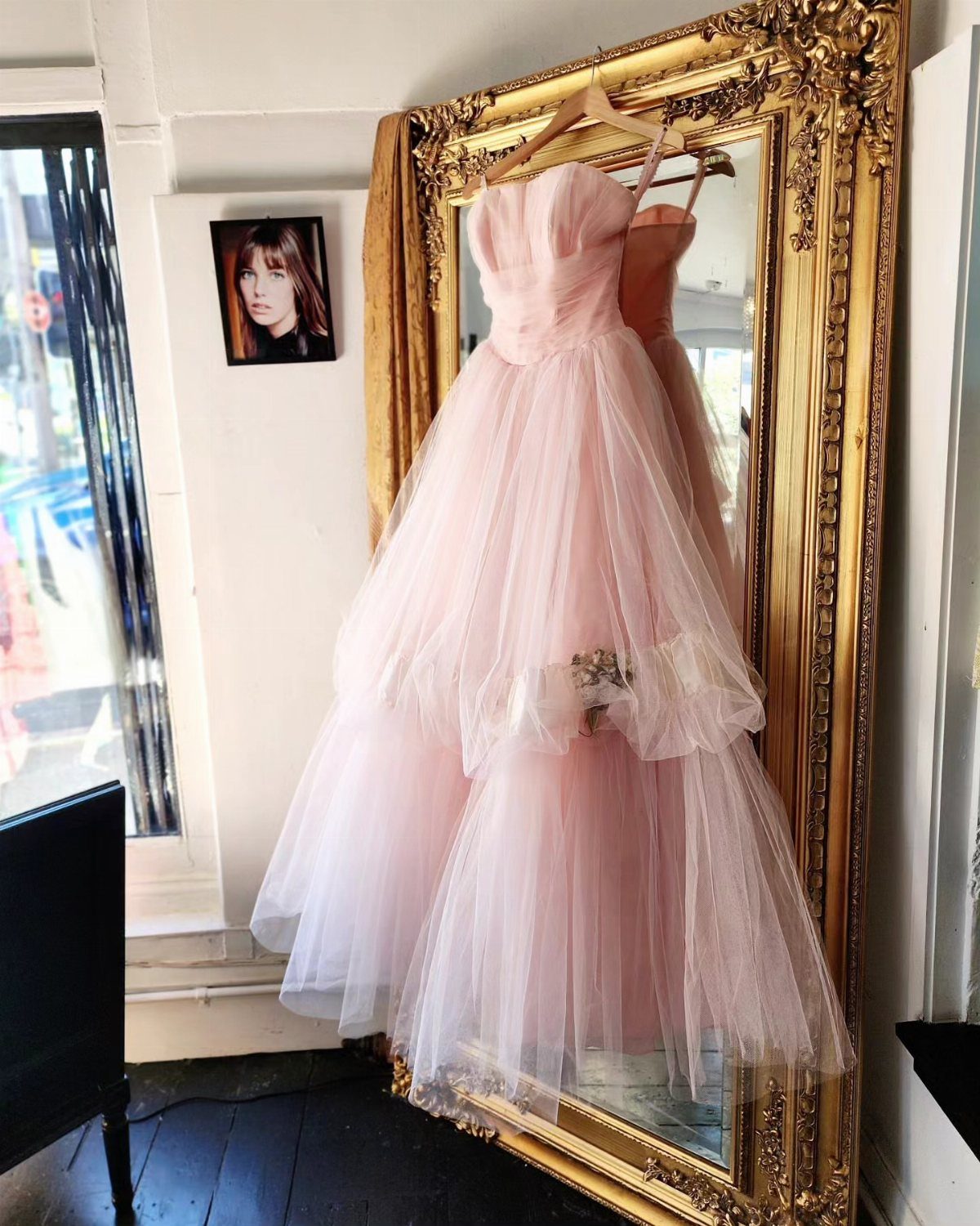 Light pink tiered tulle gown hanging before an ornate gold mirror.