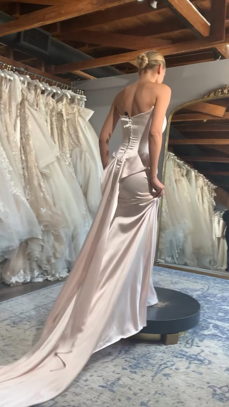 Woman wearing a strapless blush pink satin gown with a long train in a boutique.