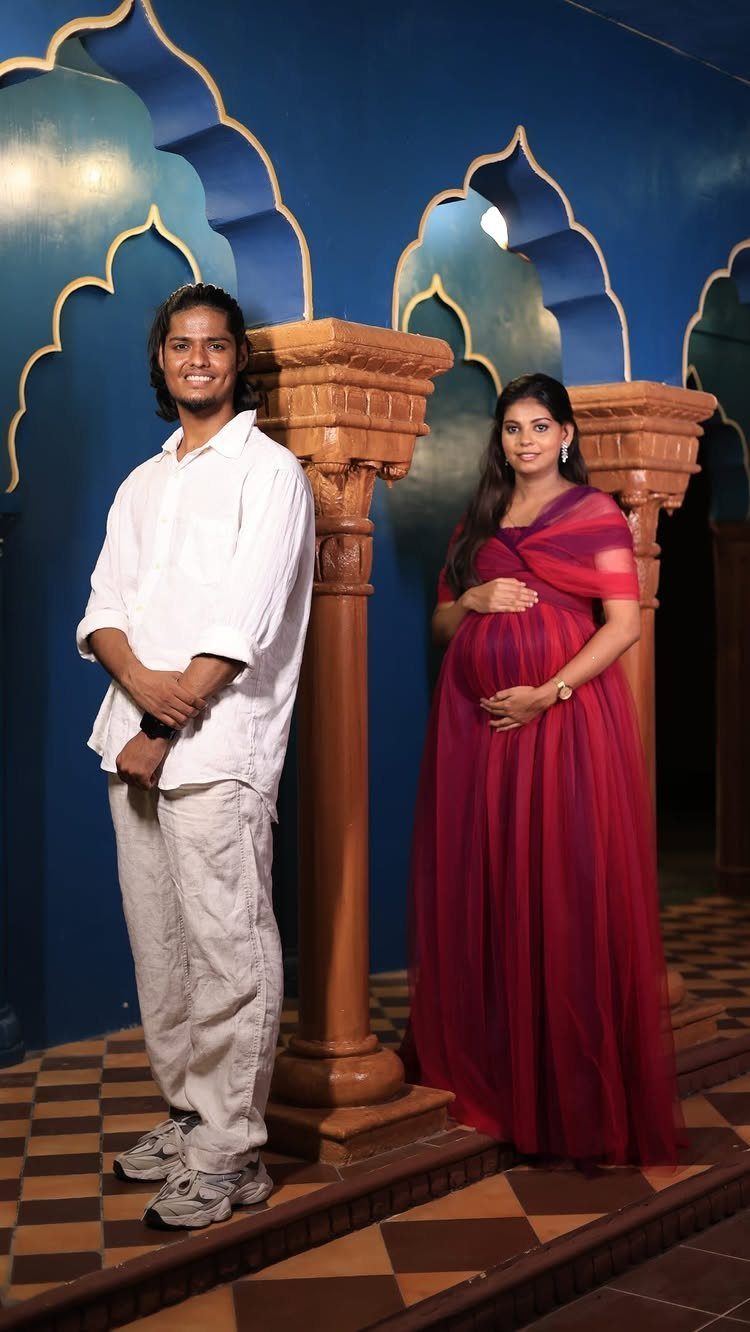 Pregnant woman in red gown and man in white posing by blue walls.