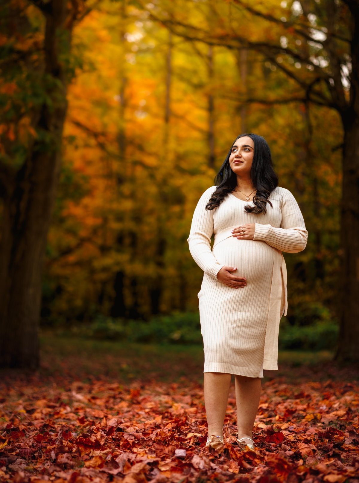 Pregnant woman in a cream knit dress standing among autumn forest leaves.