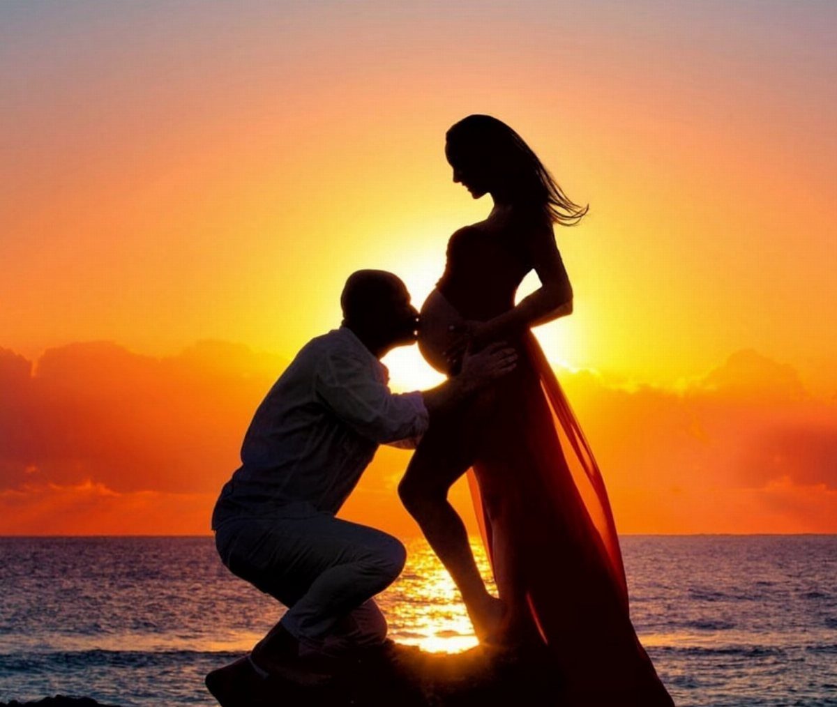 Pregnant woman in floor-length dress and man silhouetted against a sunset beach.