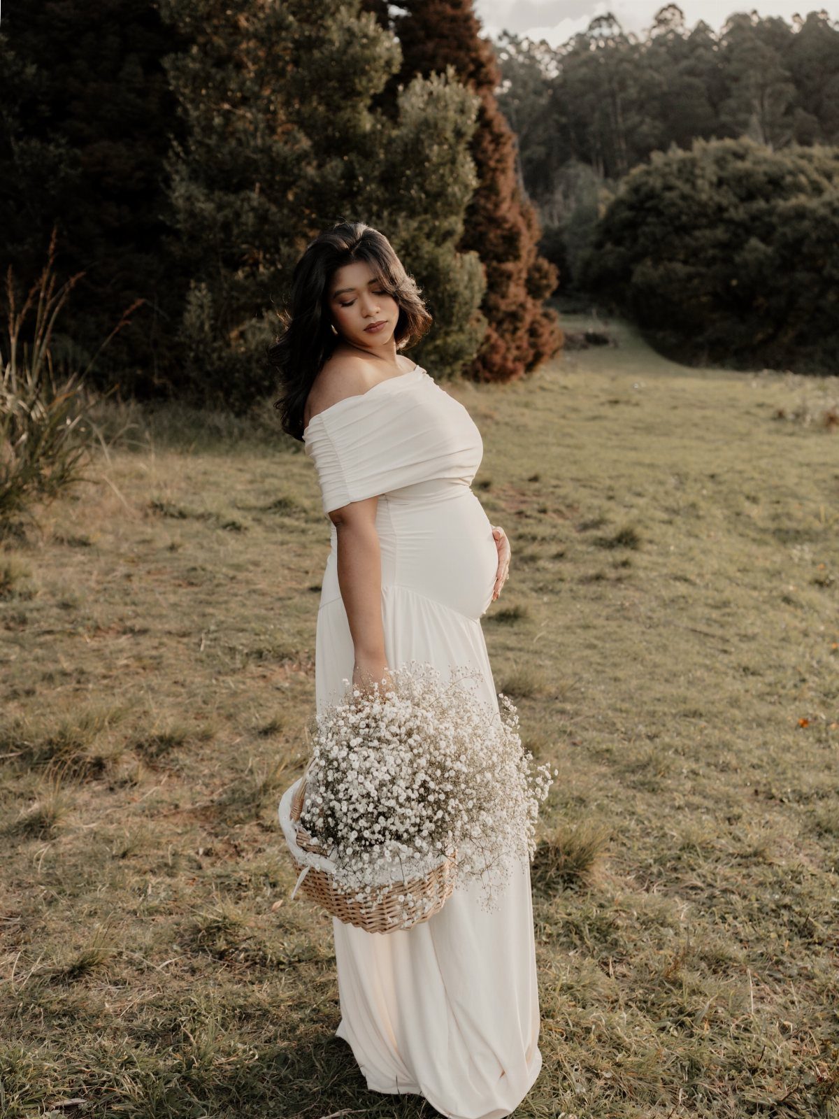 Pregnant woman in a long white off-the-shoulder dress holding a basket of flowers.