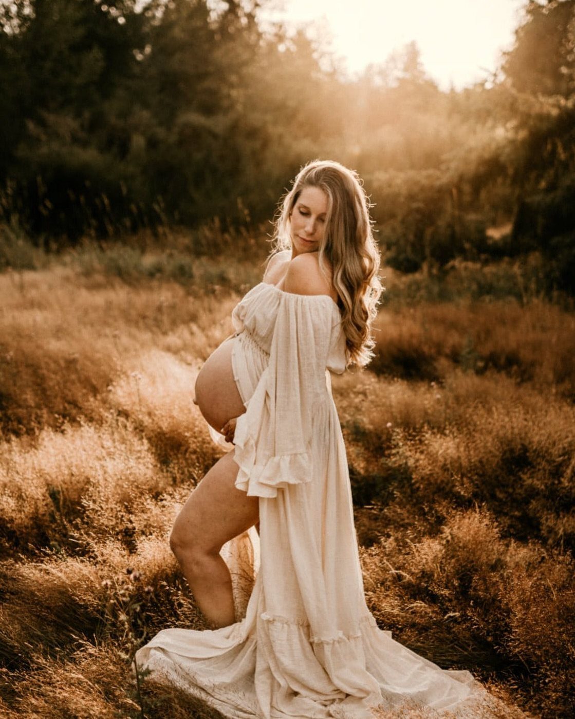 Pregnant woman in a white ruffle dress posing in a golden grassy field.