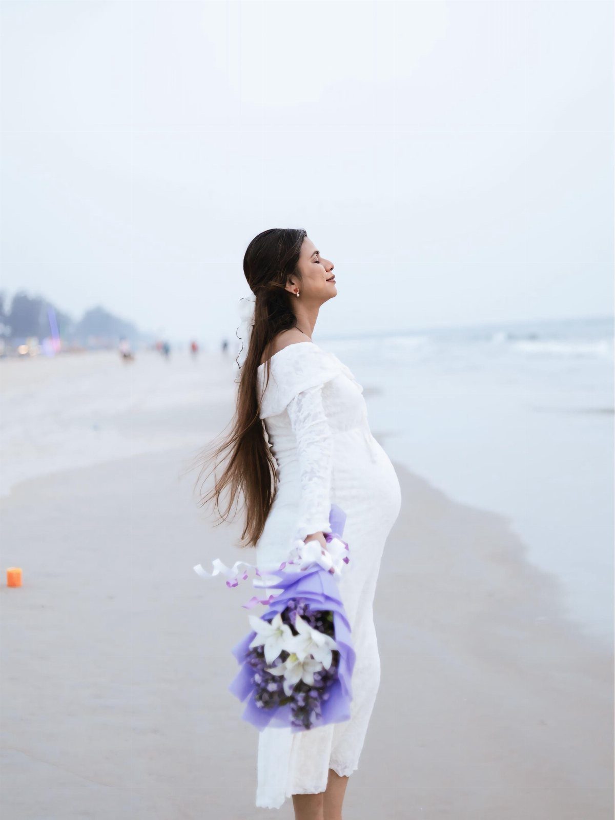Pregnant woman in white lace dress holding purple flowers on a beach