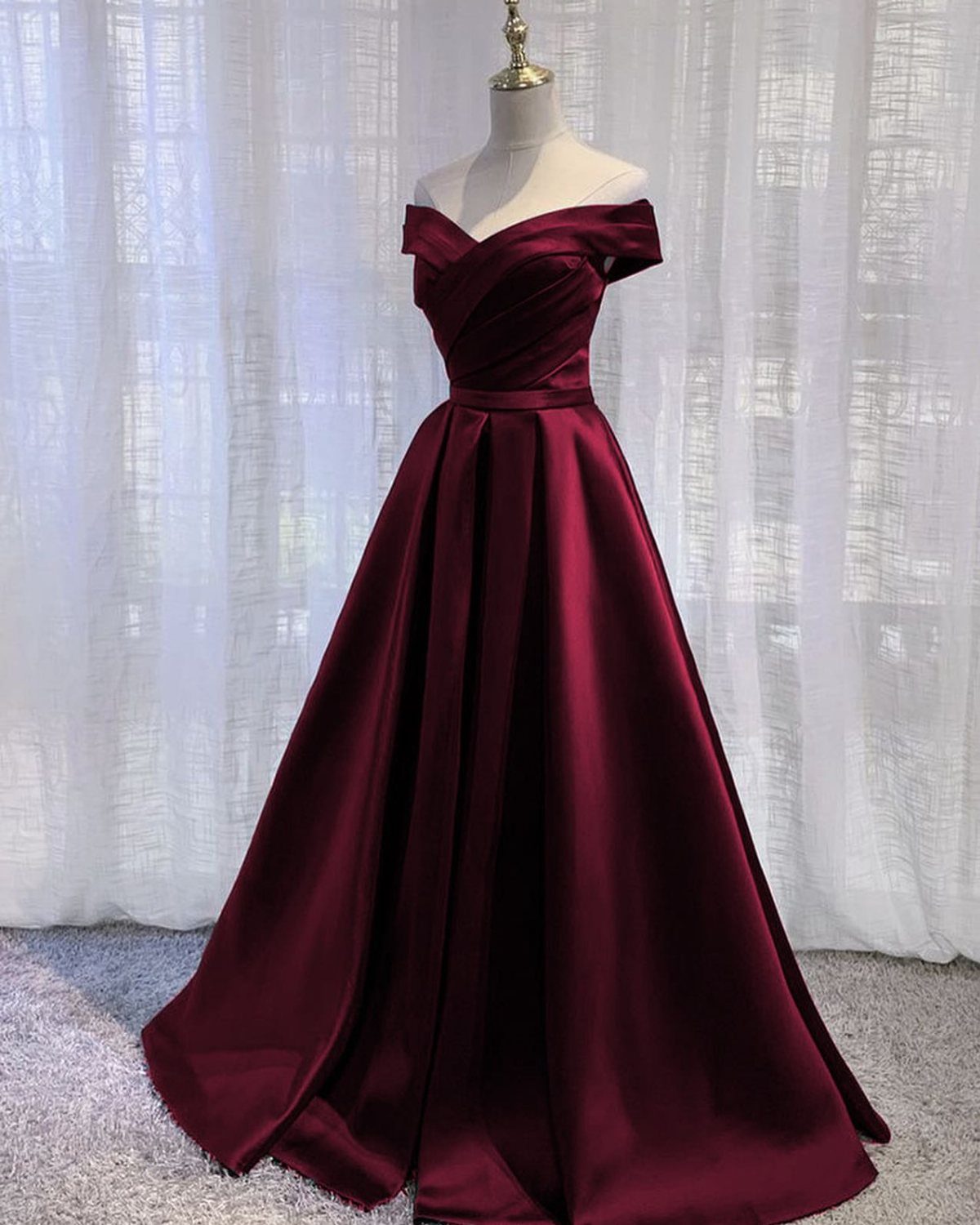 Burgundy satin off-the-shoulder prom gown on a mannequin against white curtains