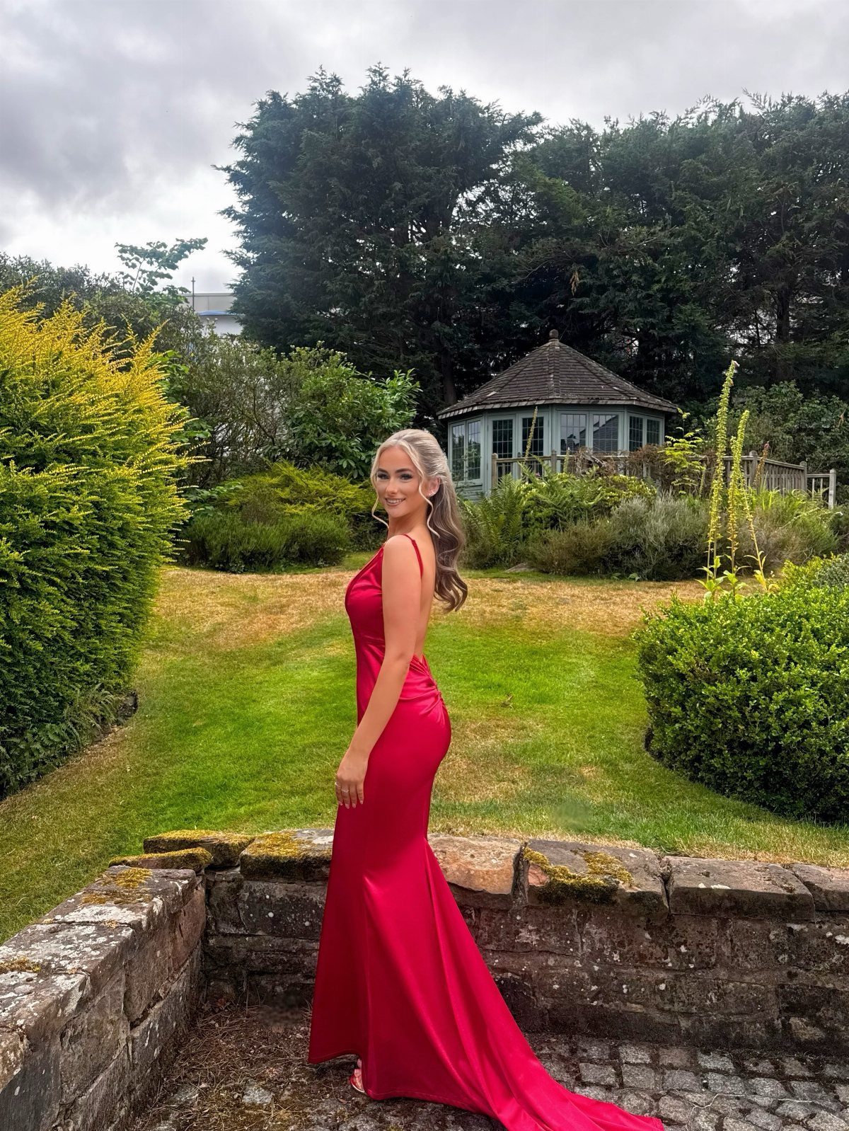 Woman in a simple red silk floor-length gown posing in a lush green garden.