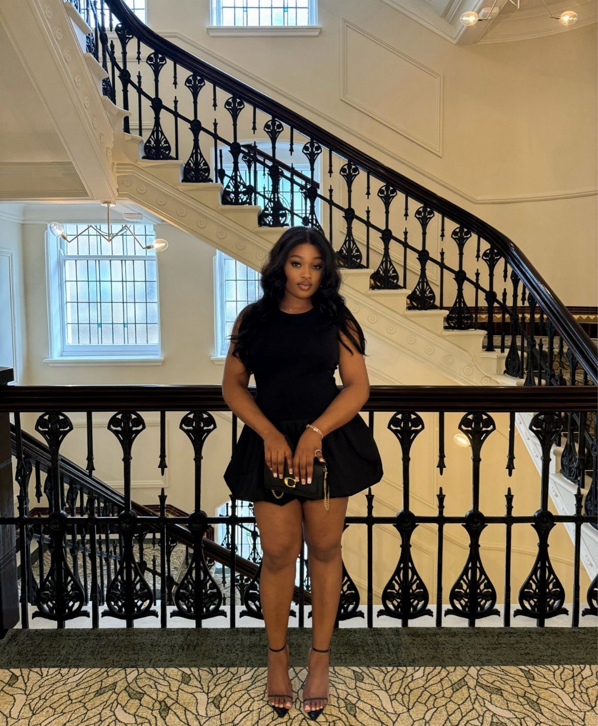 Woman in a short sleeveless black dress standing by an ornate black staircase railing.