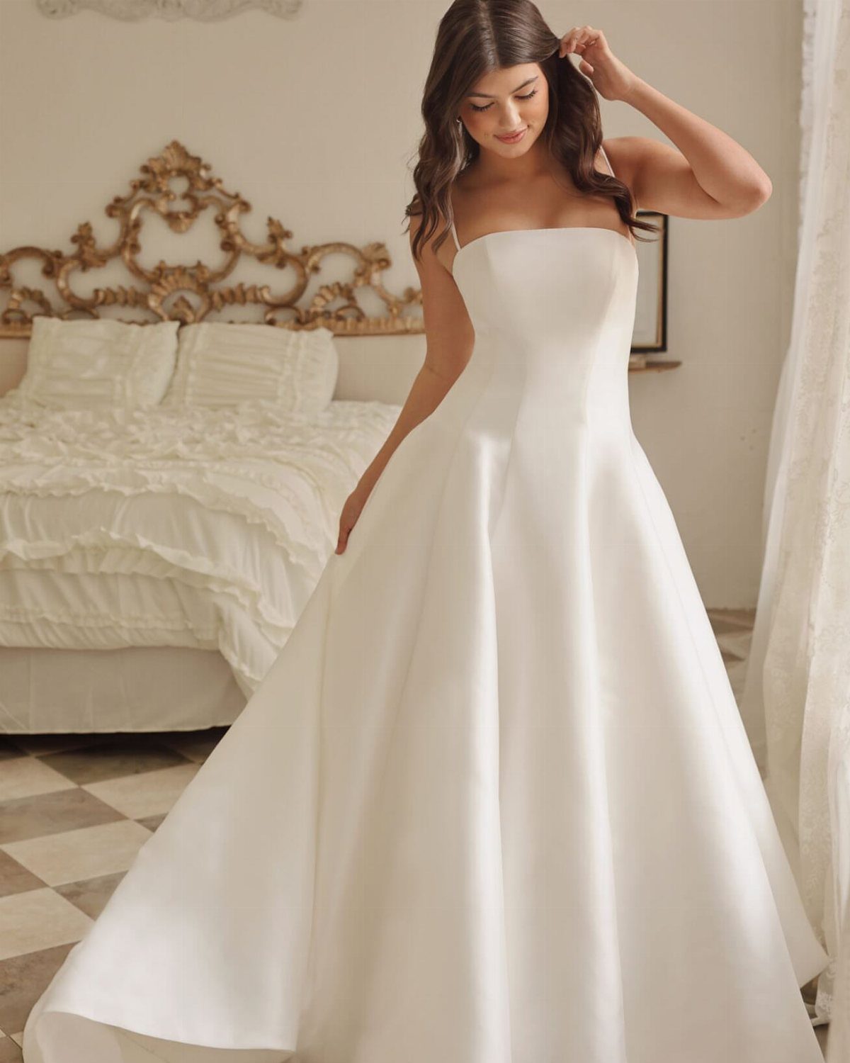 Simplistic Wedding Dress Ideas: 6 Elegant Styles to Consider 6 Woman in a strapless white satin wedding dress standing in a bright bedroom.