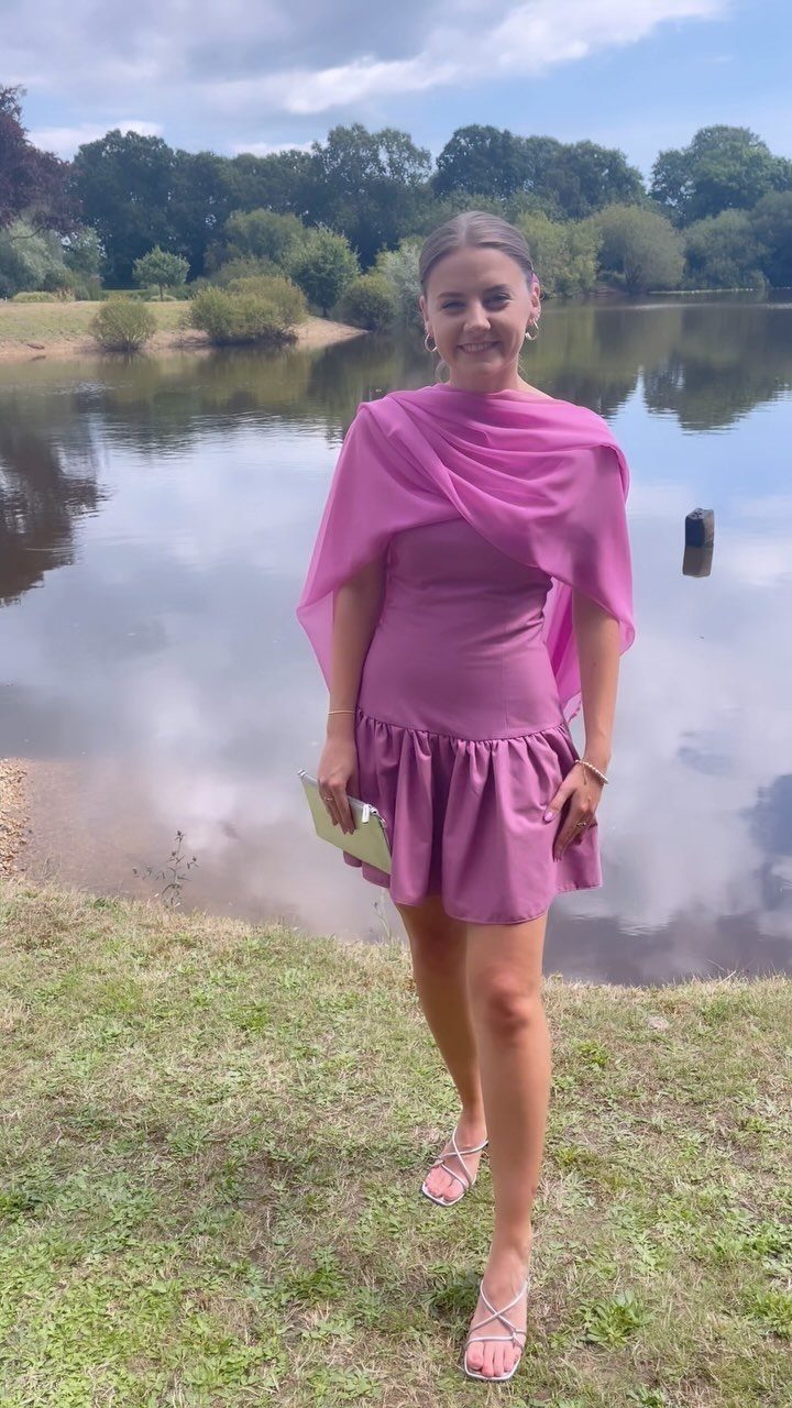 Woman in a mauve-purple cocktail dress and capelet standing by a calm lake.