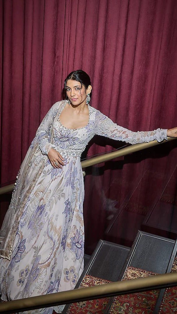 Woman in a floor-length white gown with purple floral patterns standing on stairs.