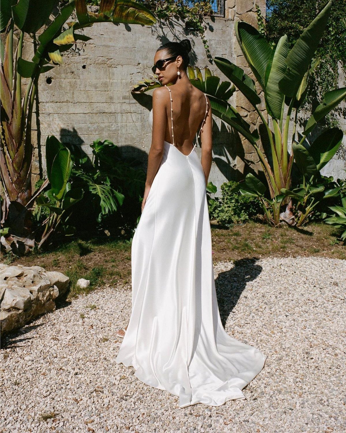 Woman in a long white satin slip dress standing outdoors by tropical plants.